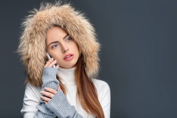beautiful woman in fur hat and winter outfit talking on smartphone, isolated on grey - Photo, Image