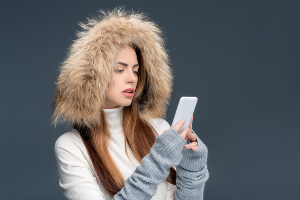 attractive woman in fur hat using smartphone, isolated on grey - Фото, изображение