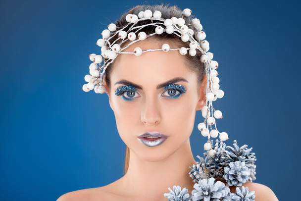 beautiful model with hair accessory, christmas pine cones, winter makeup and glitter, isolated on blue - Fotó, kép