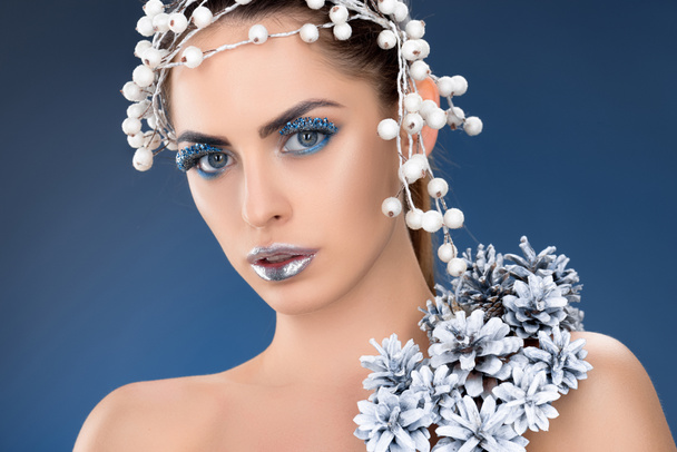 Portret van aantrekkelijke vrouw met haartoebehoren, kerst dennenappels, winter make-up en glitter, geïsoleerd op blauw - Foto, afbeelding