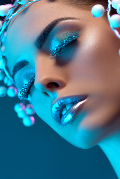 attractive woman with hair accessory, winter makeup and glitter posing for fashion shoot, isolated on blue - Photo, Image