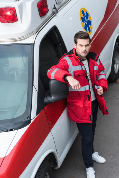 serious young male paramedic standing and leaning on ambulance - Zdjęcie, obraz