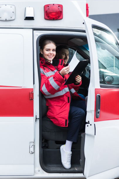 smiling paramedic sitting in ambulance and holding clipboard - Foto, imagen