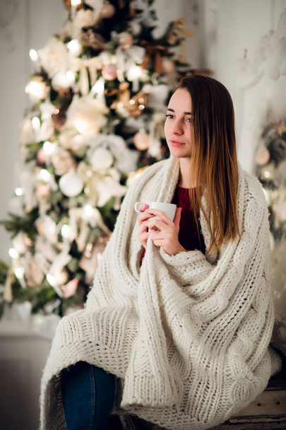 Happy young woman with cup of hot beverage near christmas tree - Фото, изображение