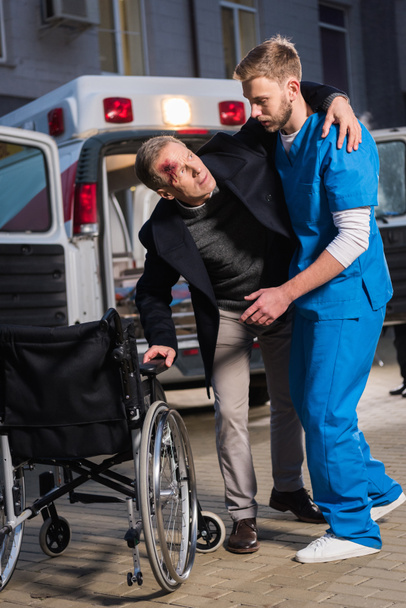 paramédico ayudando al paciente lesionado a sentarse en silla de ruedas
 - Foto, Imagen