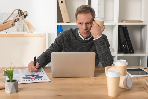 уставший бизнесмен сидит за ноутбуком с одноразовой чашкой кофе
 - Фото, изображение