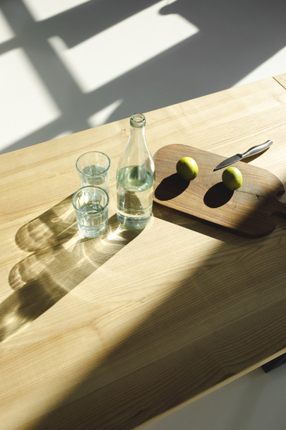 overhead view of limes on wooden board and water in glasses and bottle on wooden table - Zdjęcie, obraz
