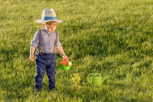 Portrait of toddler child outdoors. Rural scene with one year old baby boy wearing straw hat using watering can - Foto, Imagem