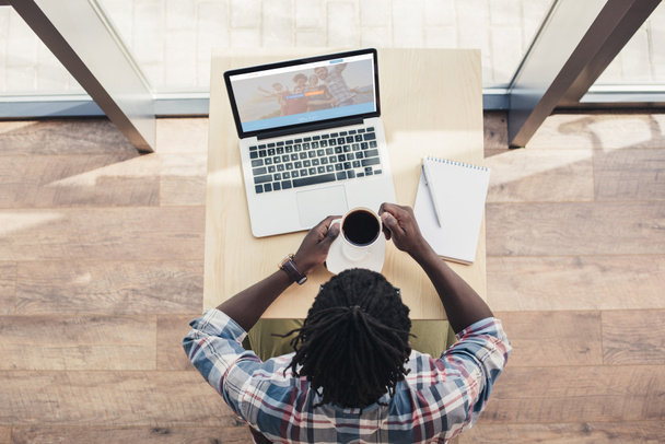 вид сверху на африканского американца, пьющего кофе и пользующегося ноутбуком с веб-сайтом по каучсерфингу
 - Фото, изображение