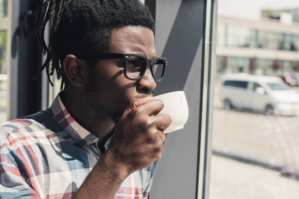 african american man drinking coffee while standing at window - Fotó, kép