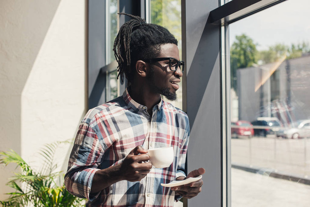 Afro-Amerikaanse man, drinken koffie terwijl staande op venster - Foto, afbeelding
