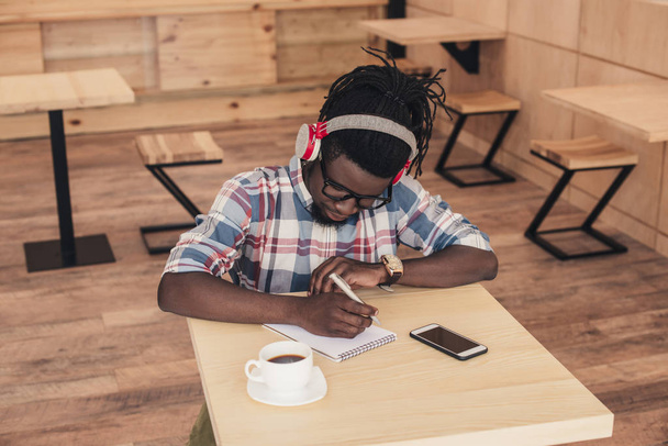 african american man listening music and writing in notepad in coffee shop  - Фото, изображение