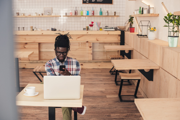 Afrikkalainen amerikkalainen mies käyttää kannettavaa tietokonetta ja älypuhelinta kahvilassa
 - Valokuva, kuva