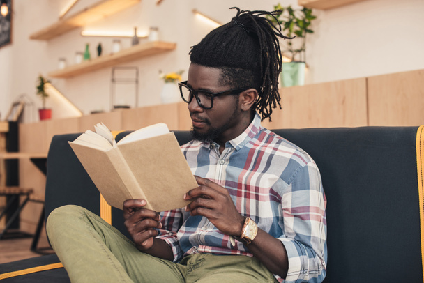 african american man reading book while sitting on sofa in cafe - Photo, Image