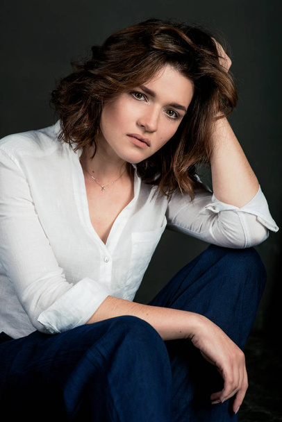 Retrato de una joven y hermosa actriz triste con pelo castaño corto en camisa blanca y pantalones azules en el estudio
 - Foto, imagen