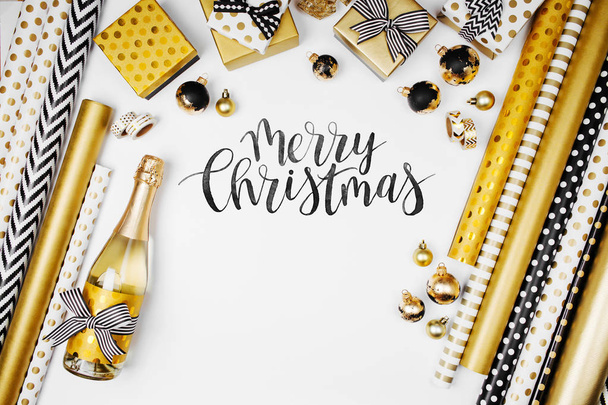 flat lay of champagne bottle, presents and golden wrapping paper rolls on white background. Merry Christmas text  - Foto, Imagen
