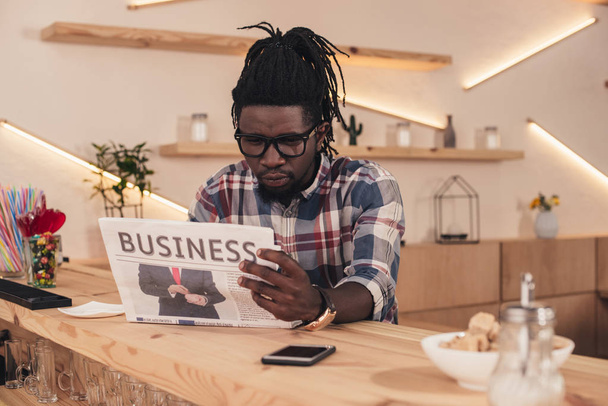 Afrikkalainen amerikkalainen mies lukee liiketoiminnan sanomalehti istuessaan baarissa laskuri älypuhelin
 - Valokuva, kuva