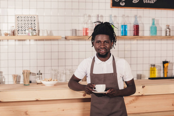 Αφρικανική αμερικανική barista στην ποδιά κρατώντας φλιτζάνι καφέ, ενώ στέκεται στην γραμμή μετρητή - Φωτογραφία, εικόνα
