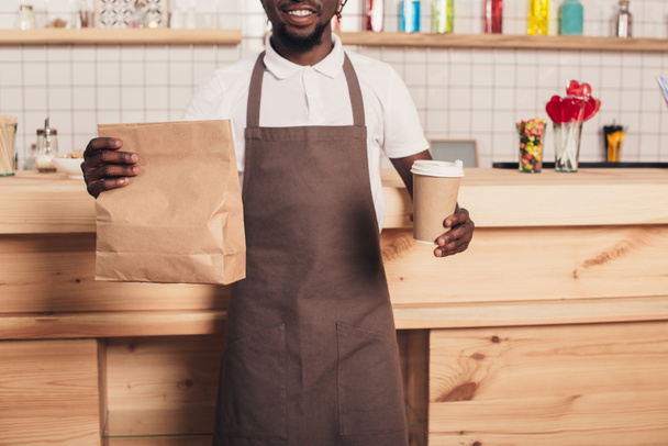 vista recortada de barista afroamericano sosteniendo taza de café desechable y paquete kraft en el mostrador de bar
 - Foto, Imagen