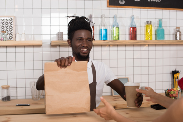 sonriendo barista afroamericano dando taza de café desechable y paquete kraft al cliente
 - Foto, imagen