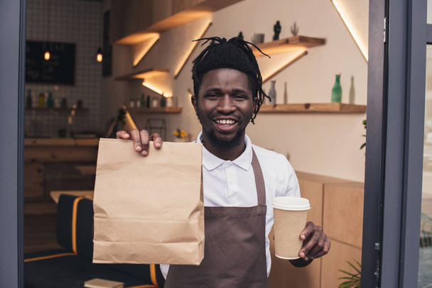 mosolyogva afro-amerikai barista gazdaság eldobható csésze kávé és a kraft csomag - Fotó, kép