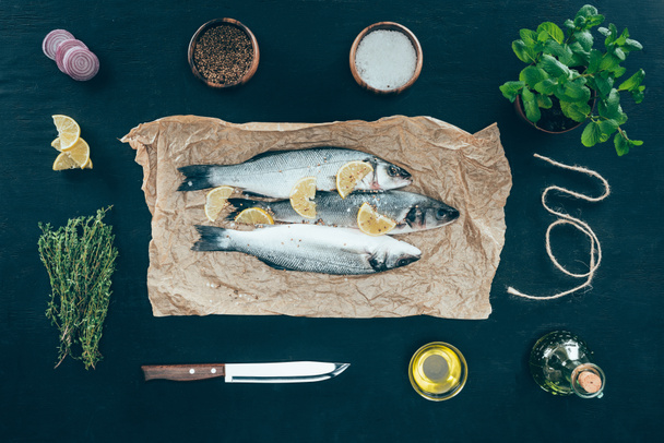 top view of gourmet fish with lemon slices on baking paper and spices on black - Foto, afbeelding