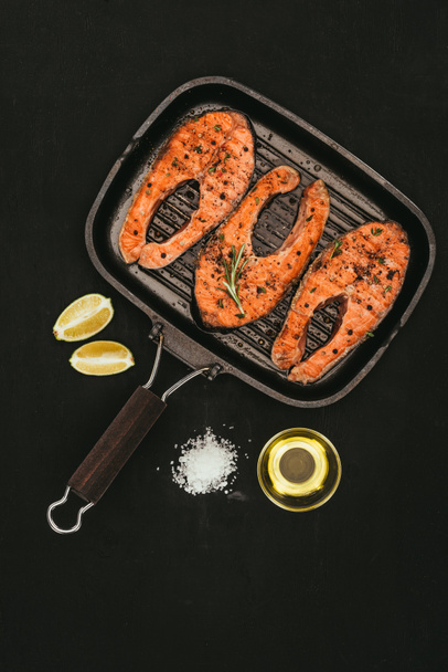 top view of salmon steaks on grill, salt, olive oil and lime slices on black - Fotó, kép