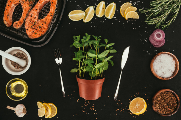 top view of mint in pot with fork and knife, salmon steaks and seasonings on black - Photo, Image