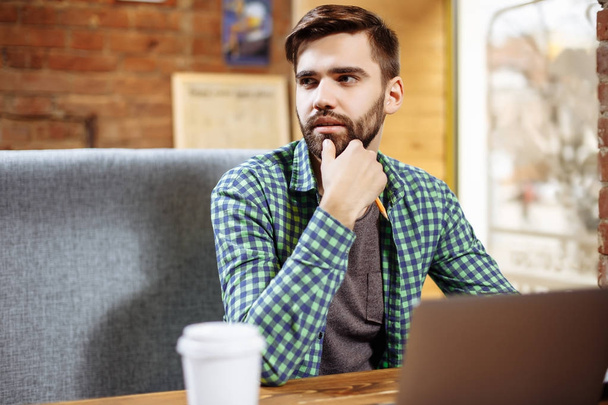 Bild eines bärtigen jungen Mannes, der in einem Café sitzt, während er einen Laptop benutzt. - Foto, Bild