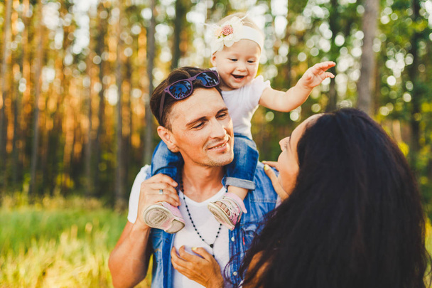 stylish young Family mom, dad and daughter one year old blonde sitting with father on shoulders playing happy and smiling, outdoors outside the city in the woods in summer at sunset. - Photo, Image