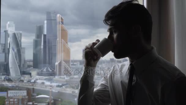 Businessman drinking coffee and looking at Moscow city towers from a window - Imágenes, Vídeo
