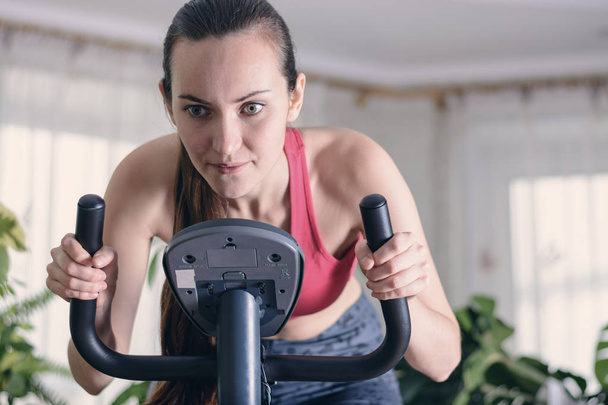 Europejska kobieta trenuje cardio trening w domu na rowerze treningowym. Koncepcja odchudzania. Zmotywowana i skupiona młoda dziewczyna - Zdjęcie, obraz