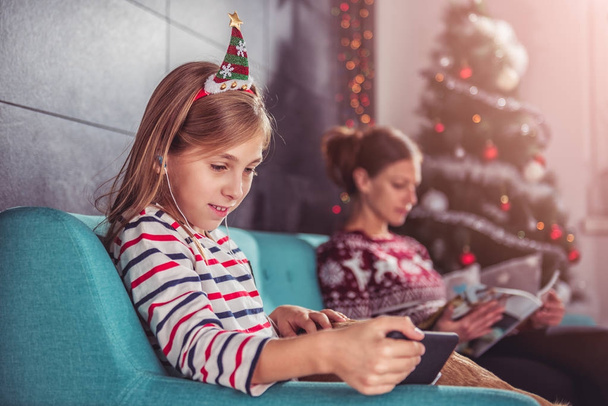 Daughter and mother relaxing on blue sofa in front of christmas tree and using smart phone and listening music - Φωτογραφία, εικόνα