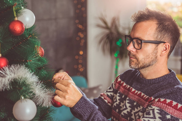 handsome man wearing blue sweater and eyeglasses decorating Christmas tree at home - Фото, изображение