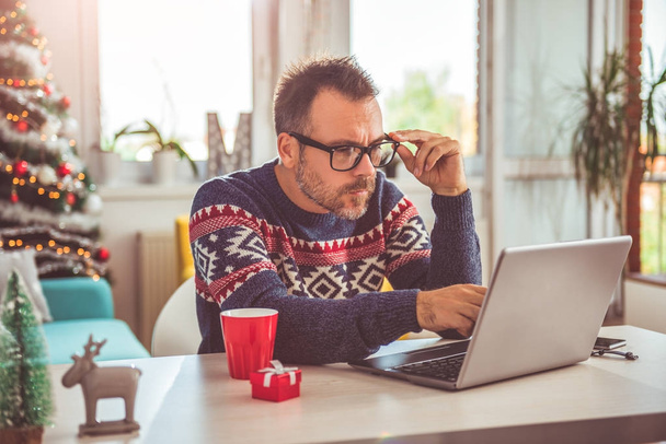 Man wearing blue sweater and eyeglasses using laptop at home with a  christmas tree in the background - Foto, Bild