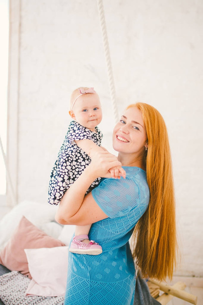 молода красива жінка з рудим довгим волоссям у блакитній сукні тримає дитину на руках один рік біля ліжка біля блондинки, на якій лежить чоловік відпочиває
 - Фото, зображення