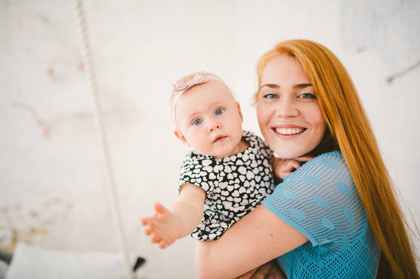 junge schöne Frau mit roten langen Haaren in einem blauen Kleid hält ein einjähriges blondes Kind auf ihren Händen in der Nähe des Bettes, auf dem ein Mann ruht - Foto, Bild
