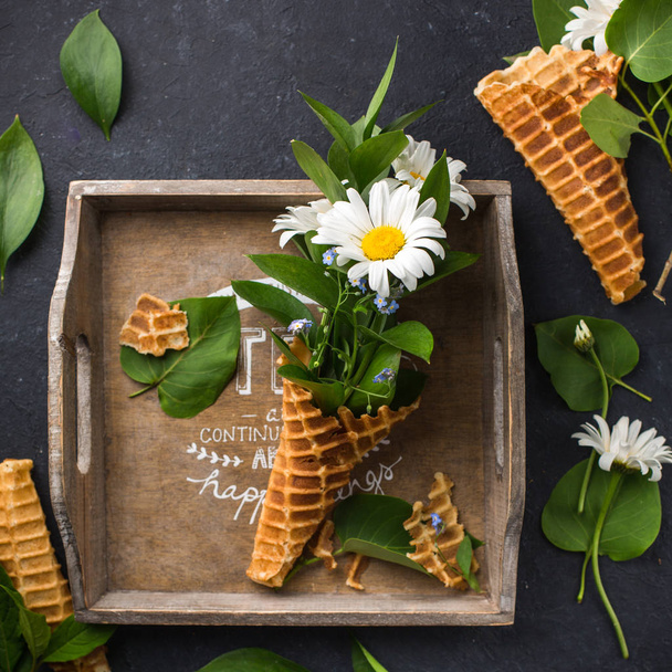 Ice cream cone with bouquet of chamomiles on a black concrete background. Fresh flowers. beautiful bouquet of wildflowers. Copy Space. Place for text. Food photo with hands. Flat lay, top view - Photo, Image