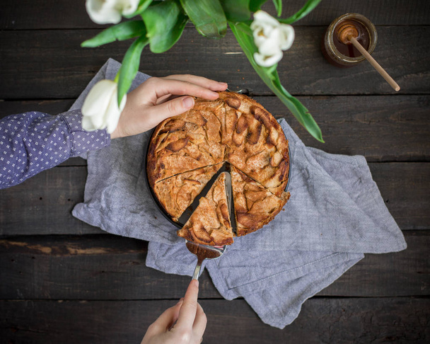 Домашний торт с цельной пшеничной мукой с яблоками на деревянном фоне. Фотография еды с руками и цветами. Полезное питание для выпечки. Плоский, вид сверху
 - Фото, изображение