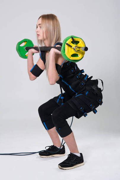 Beautiful blonde in an electric muscular suit for stimulating exercises with a barbell - 写真・画像