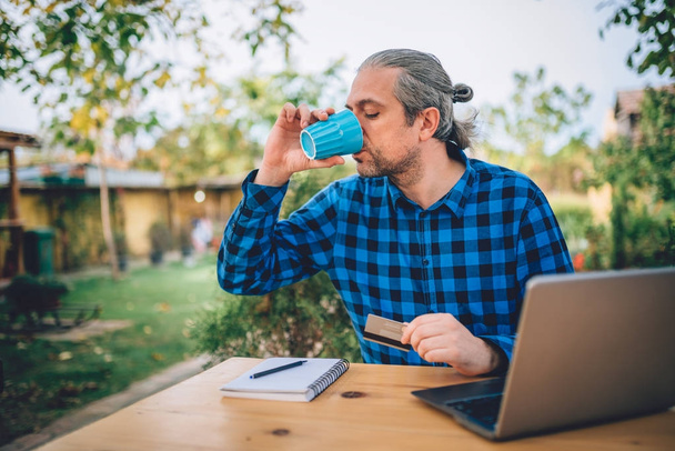 Man wearing blue plaid shirt sitting in the backyard patio, holding credit card and drinking coffee - Foto, Bild