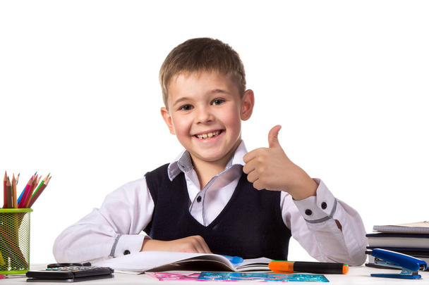 Cheerful satisfied pupil sitting at the desk with thumb up surrounded with stationery - Фото, изображение