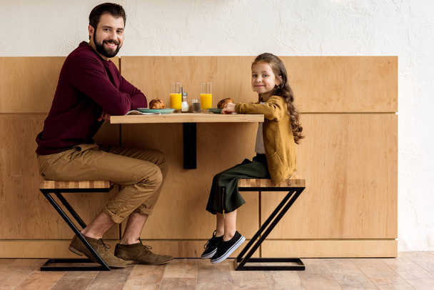 smiling daughter and father sitting at table in cafe and looking at camera - Foto, imagen