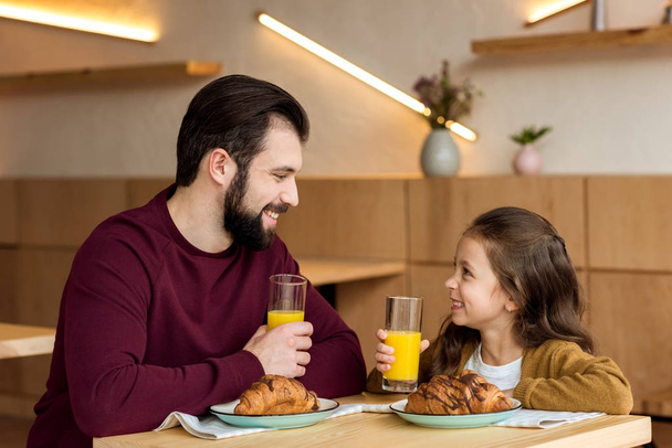 happy father and daughter holding glasses with orange juice and looking at each other  - Foto, Imagen