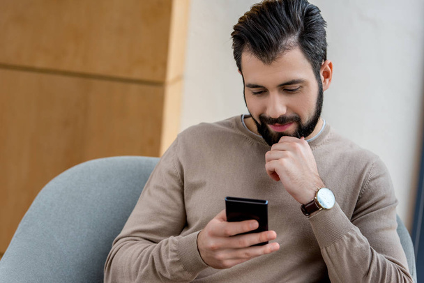 handsome man sitting in armchair and looking at smartphone - Фото, зображення