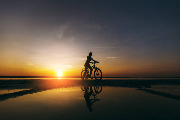 Силует спортивної дівчини в костюмі, який сидить на велосипеді у воді на заході сонця в теплий літній день. Фітнес-концепція. Небесний фон
 - Фото, зображення