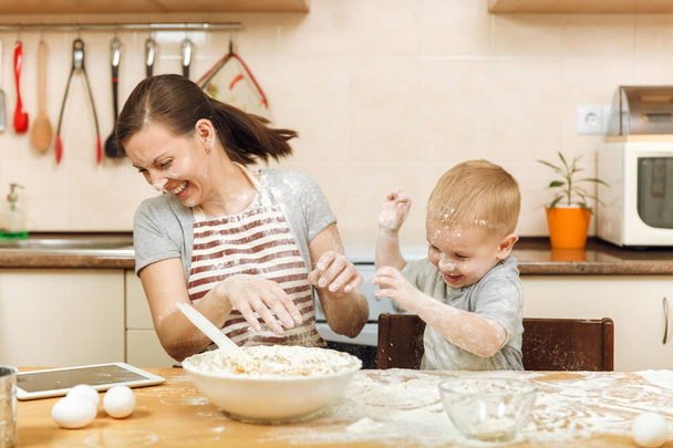 Dítě chlapec pomáhá matka vařit vánoční zázvorové sušenky v lehké kuchyni s tabletem na stole. Šťastná rodina máma 30-35 let a děti 2-3 bavit a házet mouka domů. Koncepce vztahu - Fotografie, Obrázek