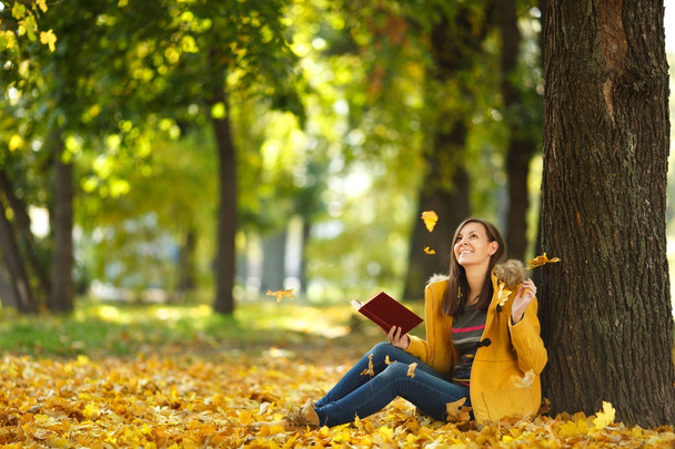 Piękny szczęśliwy uśmiechający się Brunet Kobieta w żółtym fartuchu i dżinsy, siedząc pod drzewem klonowym czerwony książkę w Upadek miasta park na ciepły dzień. Autumn golden odchodzi. Koncepcja czytania - Zdjęcie, obraz