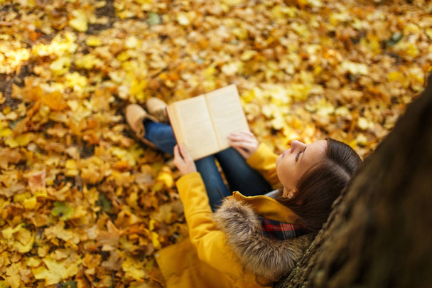 A beautiful happy smiling brown-haired woman in yellow coat and jeans sitting under the maple tree with a red book in fall city park on a warm day. Autumn golden leaves. Reading concept. Top view. - Photo, Image