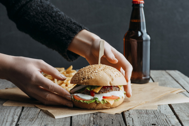 cropped view of female hands with hamburger, french fries and bottle of beer on baking paper on wooden table - Photo, Image